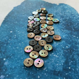 Peut inclure: Un assortiment de boutons ronds de différentes couleurs, dont irisé, marron et noir. Chaque bouton comporte deux trous pour la couture. Les boutons sont dispersés sur une surface bleue.