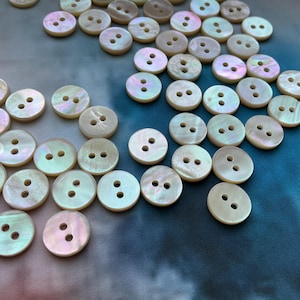 May include: A selection of iridescent, round buttons, each with two holes. The buttons have a pearlescent sheen, reflecting white, pink, and green hues. They are scattered on a blue and grey background, suggesting a craft or sewing theme.