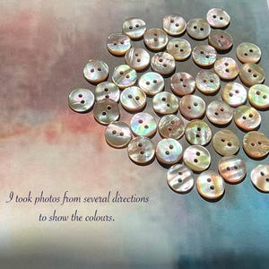 May include: A scattering of iridescent, round buttons, each with two holes. The buttons are arranged on a surface with a gradient of pink, purple, and blue hues. The image includes the text: "I took photos from several directions to show the colours."