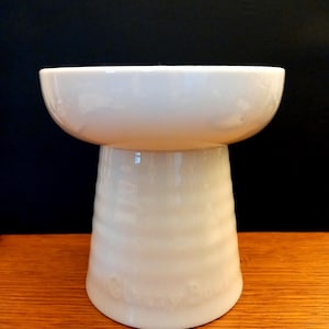 May include: A white ceramic bowl with a wide, smooth rim and a pedestal base featuring horizontal ridges. The base has the embossed text "Glassy Bowl" near the bottom. The object has a glossy finish and a uniform off-white colour throughout.