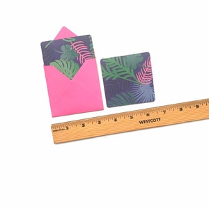 Puede incluir: Un sobre rosa con una tarjeta con estampado de hojas tropicales en su interior. Una tarjeta separada con estampado de hojas tropicales est&aacute; al lado del sobre. Una regla de madera est&aacute; debajo de las tarjetas.