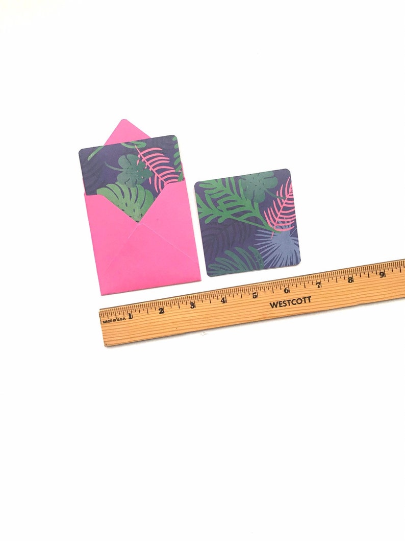 Puede incluir: Un sobre rosa con una tarjeta con estampado de hojas tropicales en su interior. Una tarjeta separada con estampado de hojas tropicales est&aacute; al lado del sobre. Una regla de madera est&aacute; debajo de las tarjetas.