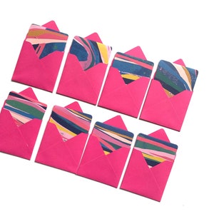 Op de afbeelding: Acht roze enveloppen met kleurrijke abstracte ontwerpen erin. De enveloppen zijn gevouwen en gerangschikt in twee rijen van vier.