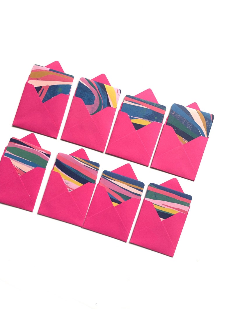 Op de afbeelding: Acht roze enveloppen met kleurrijke abstracte ontwerpen erin. De enveloppen zijn gevouwen en gerangschikt in twee rijen van vier.