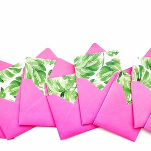 Puede incluir: Un conjunto de sobres rosas con un patrón de hojas tropicales. Los sobres están alineados en una fila, con las solapas hacia arriba.