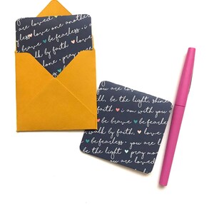 Op de afbeelding: Een set van twee kleine kaarten met inspirerende boodschappen erop gedrukt. De kaarten zijn donkerblauw met witte tekst en kleurrijke hartjes. De kaarten zitten in een gele envelop. Een roze pen is ook op de afbeelding te zien.