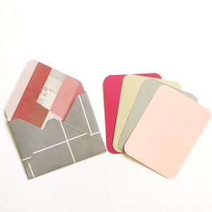 May include: A gray envelope with a white grid pattern and a pink and red striped flap. The envelope is open and a piece of paper with text is visible inside. There are five blank cards in different shades of pink, beige, and gray.