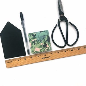 May include: A black envelope, a black pen, a ruler with the text "WESTCOTT" printed on it, a small square card with a floral pattern, and a pair of black pruning shears.