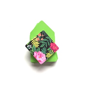 Op de afbeelding: Een stapel felgroene enveloppen met een kaart met bloemenpatroon erop. De kaart heeft roze, rode, gele en groene bloemen op een zwarte achtergrond.