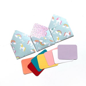 Peut inclure: Trois petites enveloppes ouvertes avec un motif de licorne et d'arc-en-ciel bleu clair, et un motif de saupoudrage rose. Plusieurs cartes carrées colorées dans les tons de rouge, orange, sarcelle, jaune, rose, blanc et lavande sont également présentes.