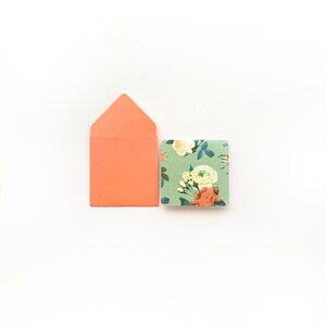 Puede incluir: Un sobre melocotón con una tarjeta con estampado floral. La tarjeta tiene un fondo verde con flores blancas y naranjas.