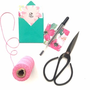 May include: A flat lay of craft supplies including a teal envelope with a floral card inside, a pink ball of twine, a black pen, and black scissors with floral patterned paper.