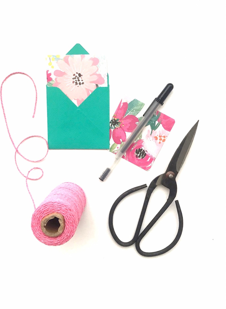 May include: A flat lay of craft supplies including a teal envelope with a floral card inside, a pink ball of twine, a black pen, and black scissors with floral patterned paper.