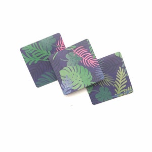 Puede incluir: Tres posavasos cuadrados con un patr&oacute;n de hojas tropicales. Los posavasos son de color azul oscuro con hojas verdes, rosas y azul claro.