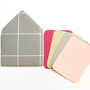 May include: A set of five blank cards in various colors, including pink, gray, beige, and red. The cards are arranged in a fan shape and are next to a gray envelope with a white grid pattern.