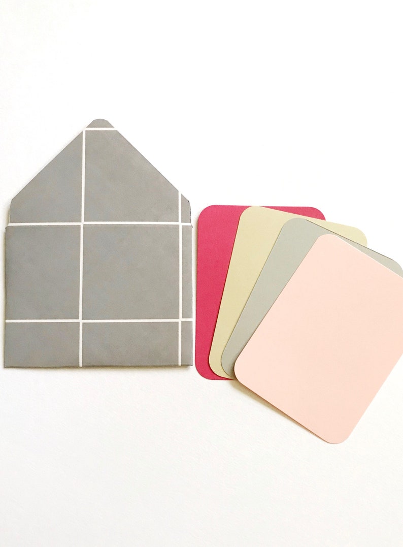 May include: A set of five blank cards in various colors, including pink, gray, beige, and red. The cards are arranged in a fan shape and are next to a gray envelope with a white grid pattern.