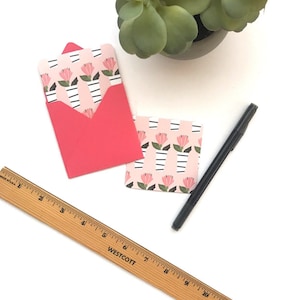 May include: A set of two pink and white floral patterned note cards with matching red envelopes. The cards are on a white surface with a wooden ruler and a black marker.