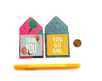May include: Two small envelopes with floral and geometric patterns. The envelopes contain cards with the text "Thank You" and "You Go Girl". A yellow marker and a penny are also included in the image.