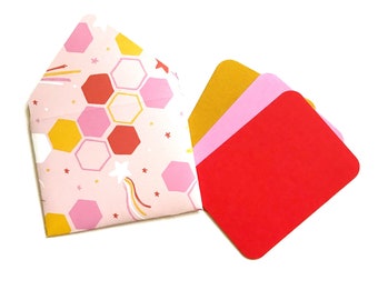 May include: A set of three blank greeting cards with envelopes. The envelope is pink with a geometric pattern of yellow and red hexagons and stars. The cards are red, pink, and yellow.