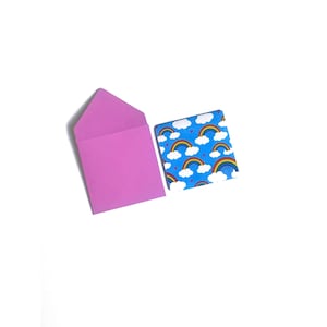 Peut inclure: Une enveloppe rose avec une carte sur fond bleu avec un motif répété d'arcs-en-ciel et de nuages blancs avec des cœurs roses.