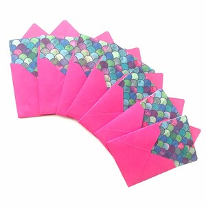 Pode incluir: Um conjunto de oito envelopes cor-de-rosa com um padrão de escamas de sereia na frente. As escamas são em tons de azul, verde e roxo.
