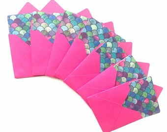 Op de afbeelding: Een set van acht roze enveloppen met een zeemeermin-schubbenpatroon aan de voorkant. De schubben zijn in tinten blauw, groen en paars.