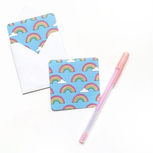 Peut inclure: Une carte bleue avec un motif arc-en-ciel et nuage, une enveloppe blanche et un stylo rose sur une surface blanche.