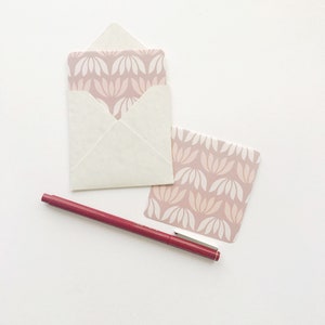 May include: A white envelope with a pink and white floral pattern on the inside. A pink and white floral patterned card is next to the envelope. A red pen is also in the image.
