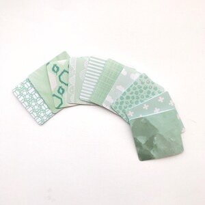 Puede incluir: Un conjunto de diez cuadrados de papel verde con patrones. Los cuadrados están dispuestos en semicírculo con el lado estampado hacia afuera. Los patrones incluyen formas geométricas, rayas y lunares.