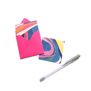 Op de afbeelding: Een roze envelop met een kaart met een kleurrijk abstract ontwerp erin. Een zilveren pen is ook inbegrepen.