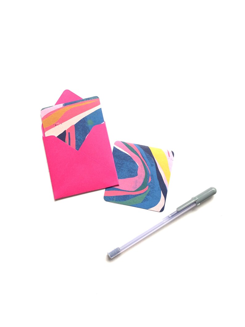Op de afbeelding: Een roze envelop met een kaart met een kleurrijk abstract ontwerp erin. Een zilveren pen is ook inbegrepen.