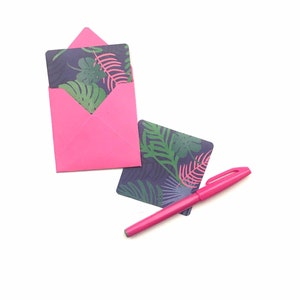 Puede incluir: Un sobre rosa con una tarjeta con estampado de hojas tropicales en su interior. Tambi&eacute;n se muestra una pluma rosa y una segunda tarjeta con estampado de hojas tropicales.