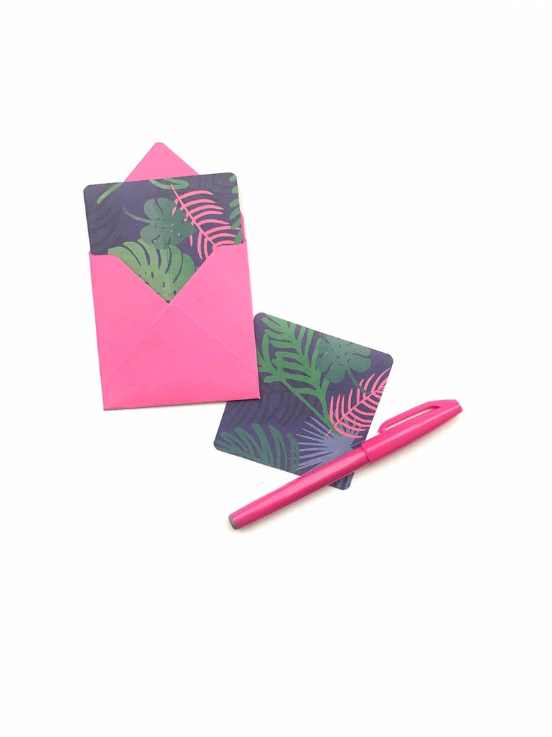 Puede incluir: Un sobre rosa con una tarjeta con estampado de hojas tropicales en su interior. Tambi&eacute;n se muestra una pluma rosa y una segunda tarjeta con estampado de hojas tropicales.