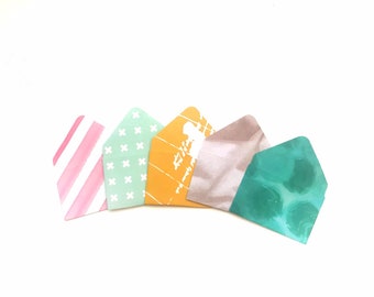 May include: Five colorful patterned envelopes with different designs. The envelopes are arranged in a semi-circle with the pink and white striped envelope on the left, followed by a green envelope with white crosses, a yellow envelope with a white design, a brown envelope with a white design, and a teal envelope with a white design.