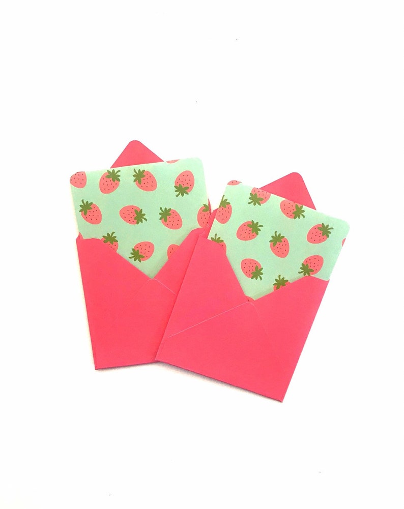 Pode incluir: Dois envelopes cor-de-rosa com um padr&atilde;o de morangos verdes e brancos na frente.