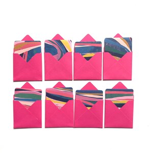 Op de afbeelding: Zes roze enveloppen met kleurrijke, abstracte patronen op de kaarten erin. De enveloppen zijn gerangschikt in twee rijen van drie.