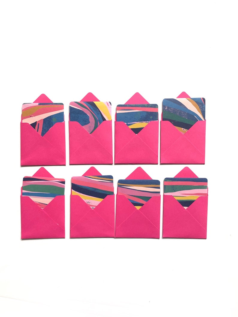 Op de afbeelding: Zes roze enveloppen met kleurrijke, abstracte patronen op de kaarten erin. De enveloppen zijn gerangschikt in twee rijen van drie.