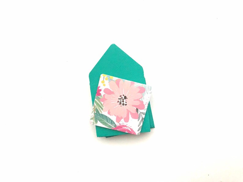 May include: A stack of teal envelopes with a floral patterned card on top. The card features a pink flower with a dark center and green leaves.
