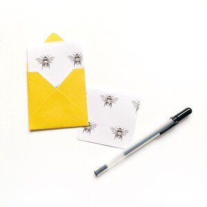 May include: A yellow envelope with a white card inside. The card has a repeating pattern of black and white bee illustrations. A black pen is lying on the white surface.