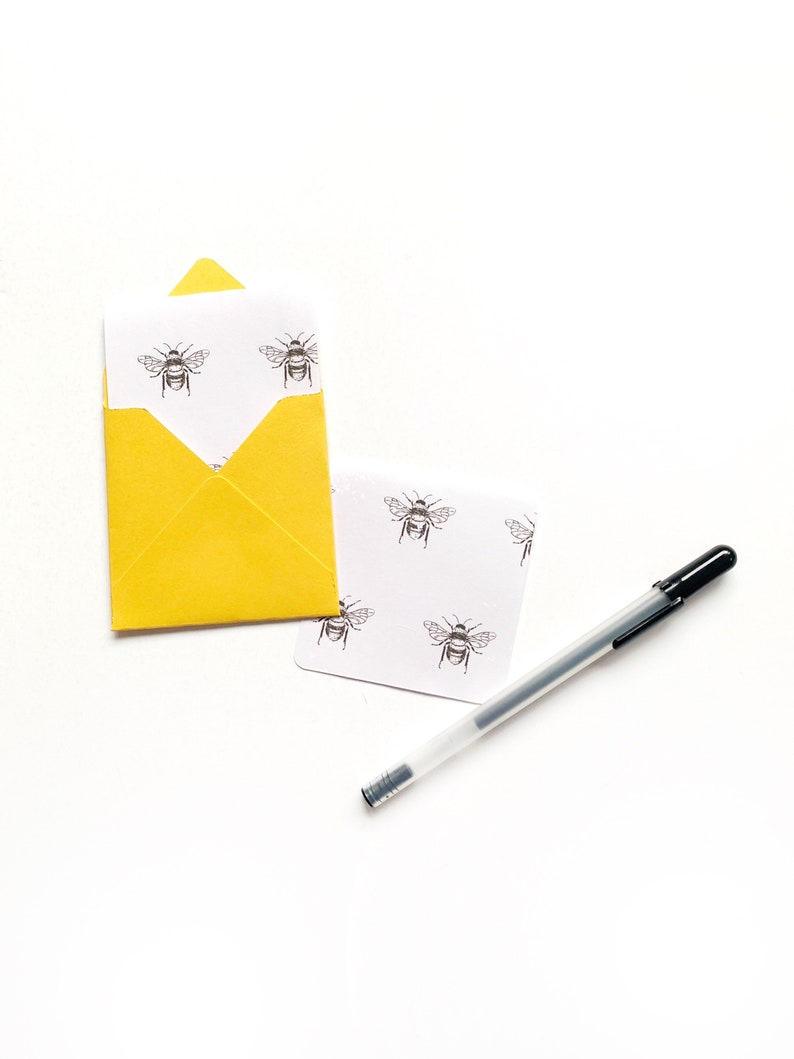 May include: A yellow envelope with a white card inside. The card has a repeating pattern of black and white bee illustrations. A black pen is lying on the white surface.