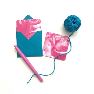May include: A teal blue envelope with a pink floral card inside, a pink pen, and a ball of teal blue yarn.