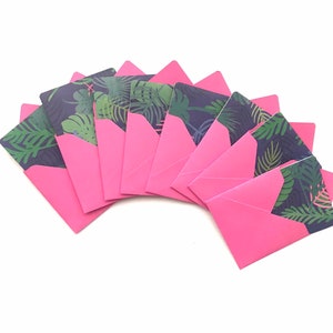 Puede incluir: Un conjunto de ocho sobres rosas con un patr&oacute;n de hojas tropicales en el frente. Los sobres est&aacute;n dispuestos en forma de abanico.