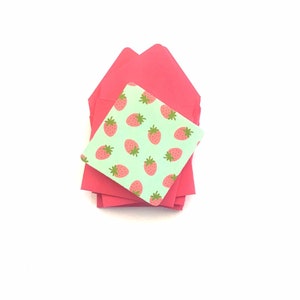 Pode incluir: Uma pilha de envelopes cor-de-rosa com um pequeno envelope verde com um padr&atilde;o de morangos no topo.