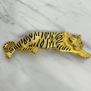 May include: A gold-toned tiger brooch with black stripes and details. The brooch is in a reclining pose, with its paws extended. The tiger's face has detailed features, including eyes and whiskers. The brooch is likely made of metal.