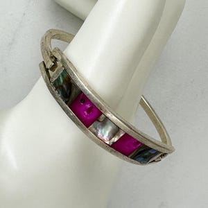 May include: A silver-tone hinged bangle bracelet with a row of alternating pink and iridescent shell squares. The bracelet is shown on a white surface.
