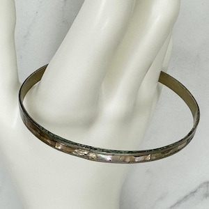 Op de afbeelding: Een dunne, zilverkleurige armband met ingelegde iriserende schelpsegmenten. De armband wordt getoond op een wit oppervlak, waarbij het delicate ontwerp en de natuurlijke kleurvariaties worden benadrukt. Een eenvoudig, elegant sieraad.