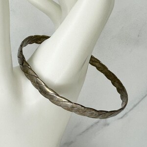 May include: A silver-toned braided bangle bracelet. The bracelet has a woven design and is displayed on a white surface. The jewelry piece is circular and appears to be made of metal.
