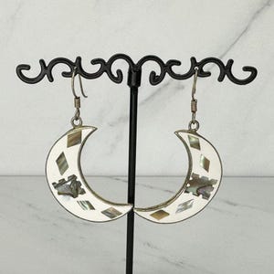 May include: A pair of crescent moon-shaped earrings featuring a silver-toned metal frame. The surface of each earring is inlaid with white material and small, iridescent, diamond-shaped pieces of shell, suspended from silver-toned metal hooks.