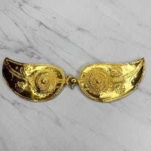 Pode incluir: Um sutiã decorativo dourado com um design ornamentado. O sutiã apresenta um fecho central e duas taças em forma de asa com intrincados padrões florais e circulares. A superfície tem um acabamento brilhante e reflexivo.