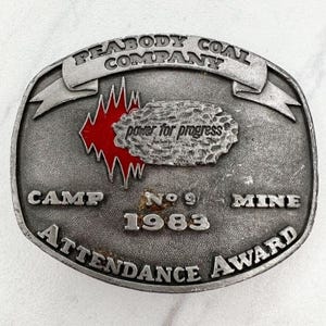 Op de afbeelding: Ovale zilverkleurige metalen riemgesp. De gesp heeft de tekst "PEABODY COAL COMPANY" bovenaan en "CAMP No 9 MINE 1983 ATTENDANCE AWARD" onderaan. Een rood-witte afbeelding en de woorden "power for progress" staan in het midden.